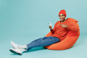 Full body young smiling satisfied fun happy african american man in orange shirt hat sit in bag chair use hold point finger on mobile cell phone isolated on plain pastel light blue background studio.