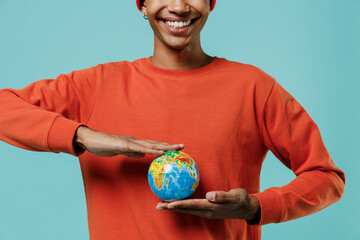 Cropped close up young smiling happy african american man in orange shirt hat hold in palms Earth world globe isolated on plain pastel light blue background studio portrait. People lifestyle concept.