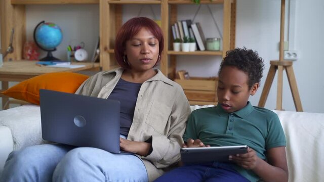 Young woman working with laptop and boy playing with tablet, sitting on couch in apartment spbd. Front view of mom does job and uses computer, son holds gadget in hands and sits on sofa in light