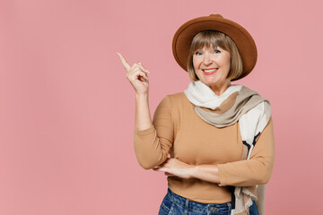 Traveler tourist mature elderly senior lady woman 55 years old wears brown shirt hat scarf pointing on workspace area copy space mock up isolated on plain pastel light pink background studio portrait.