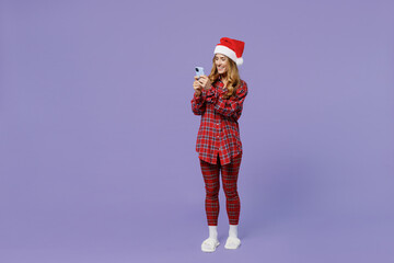 Full size body length young Santa woman 30s wears shirt Christmas hat hold in hand use mobile cell phone isolated on pastel light violet background studio portrait. Happy New Year celebration concept.