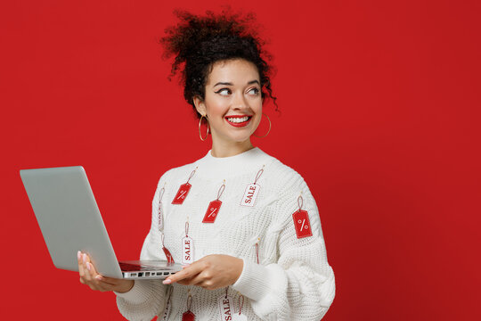 Young smiling happy female costumer woman 20s in white knitted sweater with tags sale in store showroom look aside hold use work on laptop pc computer isolated on plain red background studio portrait.