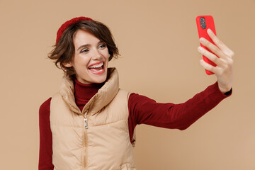 Naklejka premium Young woman 20s wears red turtleneck vest beret doing selfie shot on mobile cell phone post photo on social network isolated on plain pastel beige background studio portrait. People lifestyle concept.