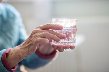 A glass of water in the wrinkled hands of an elderly woman, grandmother drinks water, health care, the benefits of drinking water for an old man.
