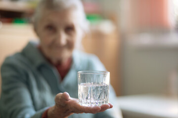 An elderly woman holds a glass glass with water in her hand, health care, grandmother drinks water for health, retirees at home during quarantine.
