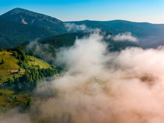 A thin morning fog covers the Ukrainian mountains. Green grass on the slopes of the mountains. A curly thin fog spreads over the mountains. Aerial drone view.