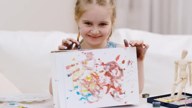Little girl shows her drawing and holding picture with hands smeared with multicolored paint