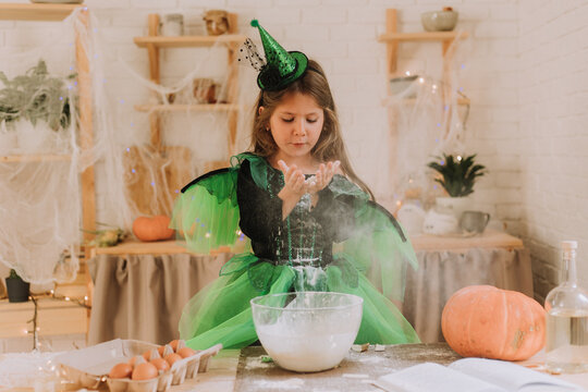 cute little girl in a green Halloween costume of a witch or fairy prepares a pumpkin pie, barm brack in a festive decorated kitchen. space for text. High quality photo