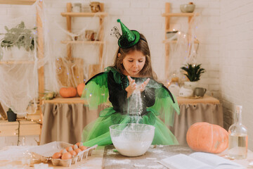 cute little girl in a green Halloween costume of a witch or fairy prepares a pumpkin pie, barm brack in a festive decorated kitchen. space for text. High quality photo