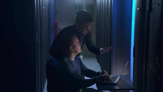 Two male engineers discussing project and working with laptop at dark room of data center spbas. Close-up view of young men talk and gesture with hands, use computer and inspect equipment racks or