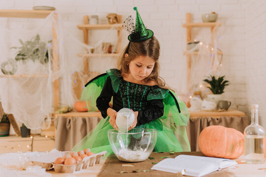 cute little girl in a green Halloween costume of a witch or fairy prepares a pumpkin pie, barm brack in a festive decorated kitchen. space for text. High quality photo