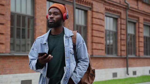 Young man listening to podcast and walking along city street with phone in hands spbas. American African guy holds smartphone and looks at screen, listens to music and walks in modern town in summer