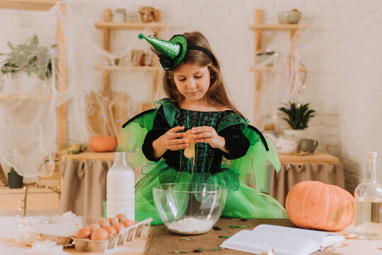 cute little girl in a green Halloween costume of a witch or fairy prepares a pumpkin pie, barm brack in a festive decorated kitchen. space for text. High quality photo