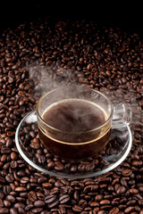 Glass of hot black coffee with glass saucer place on pile of coffee seed. hot dark coffee and vapour. close up. top view.