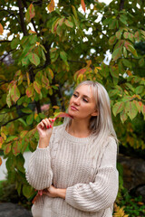 beautiful blonde woman in white knitted sweater holding red leaf in her hand, looking to side