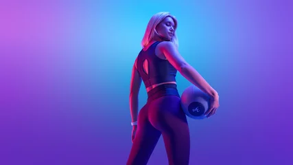 Photo sur Aluminium Fitness Confident fitness woman posing with a medicine ball. Attractive blonde sportswoman portrait holding with medicine fitness ball neon style creative light.