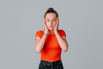 Shocked and worried brunette teen girl holding hands on head in panic, looking anxious and concerned, having big problem, standing distressed against gray background