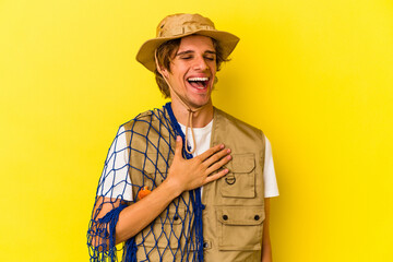 Young fisherman with makeup holding a net isolated on yellow background  laughs out loudly keeping hand on chest.