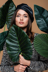 young woman in leather beret and zebra print outfit near green tropical leaves isolated on beige