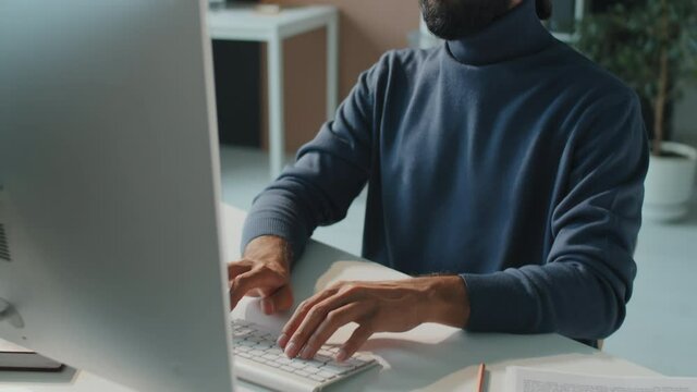 Horizontal medium shot footage of handsome young Middle Eastern man with beard on face wearing navy blue turtleneck sitting at office desk working on computer
