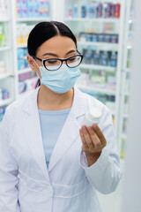 asian pharmacist in glasses and medical mask looking at bottle with pills