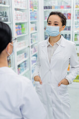 tattooed pharmacist in medical mask and white coat standing with hands in pockets near blurred colleague