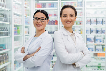 cheerful interracial pharmacists in white coats standing with crossed arms in drugstore
