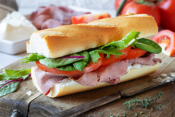 Delicious baguette sandwich with roast beef, soft cheese, tomatoes and green salad leaves on a wooden cutting board