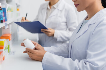 partial view of pharmacist in white coat holding bottle near blurred colleague writing on clipboard in drugstore