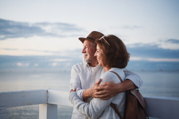 Portrait of happy senior couple in love hugging outdoors on pier by sea, looking at view, summer...