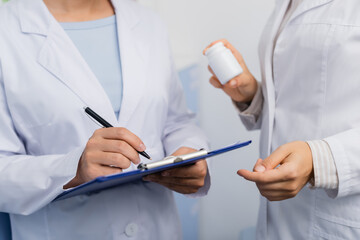 cropped view of pharmacist in white coat holding bottle near colleague writing on clipboard in drugstore