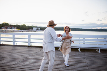 Happy senior couple in love dancing outdoors on pier by sea, looking at each other.