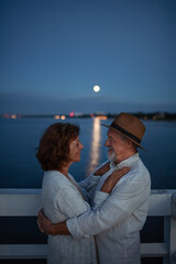 Obraz premium Happy senior couple in love hugging outdoors on pier by sea at sunset, looking at each other.