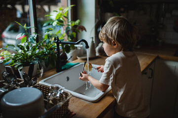 Small boy using eco friendly brush to wash dishes indoors at home, daily chores concept.