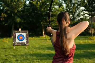 Woman with bow and arrow aiming at archery target in park, back view