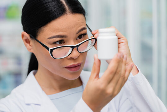 Brunette Asian Pharmacist Adjusting Glasses And Looking At Bottle With Medication