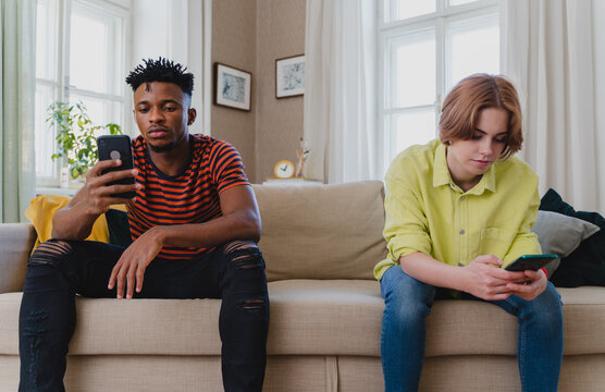Young biracial couple addicted to smartphones sitting on sofa at home, social media concept.
