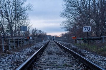 Naklejka premium Lonely empty sad front view at the iron railways heading away to the distance with sunset in the background and trees without leaves around.