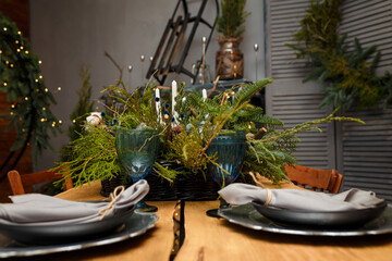 Rustic style Christmas festive table, blue glasses, plates and fresh thuja, fir branches bouquet with candles. Warm garland lights bokeh on background. Eco-friendly green decorative elements.