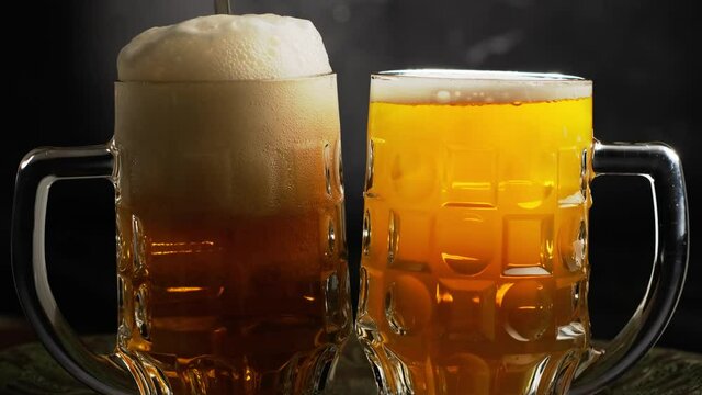 Pair of beer mugs standing on the table. Glass of beer is filled and the white foam flows down