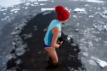 High angle view of active senior woman in swimsuit breaking ice with axe outdoors in winter, cold...