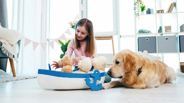 Little girl put toys to marine boat ship and playing with them. Golden retriever dog with child at home