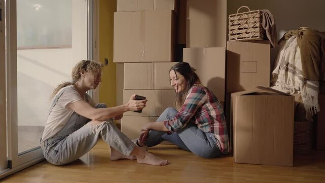 Happy women landlords tenants sit on the floor use smartphone on moving day in new house, relax between cardboard boxes discuss interior design online idea renovate home.