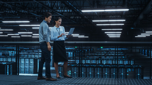 Data Center Female System Administrator and Male IT Specialist Talk, Use Laptop Computer. Information Technology Engineers work on Cyber Security Network Protection in Cloud Computing Server Farm.