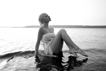 Art nude sexy blonde with short haircut is sitting in water on shore beach of lake at sunset. Wet hair and a woman body. Secluded beach holiday. Black and white
