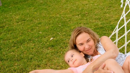 Grandmother and granddaughter, 3 years old, rest and bask in a hammock on a green meadow in their garden on a sunny summer day. Vacation concept, generational relationship, parenting, happy childhood.