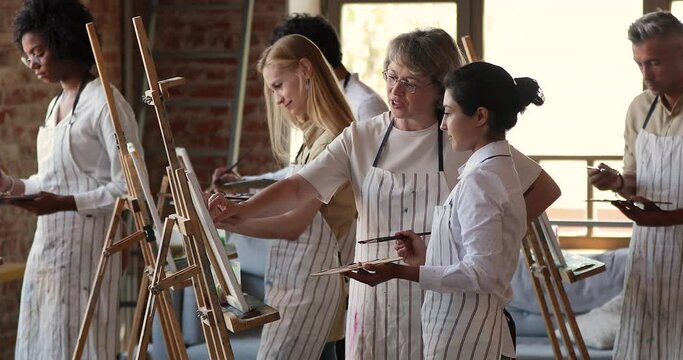 Indian student attend painting class with multi racial mates, holds palette and paintbrush drawing on easel, listens to elderly 60s female master gives advice help with technique. Art studio concept