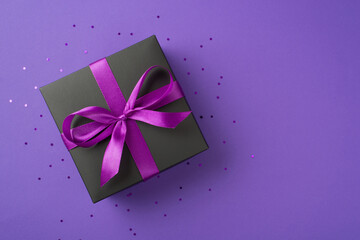 Top view photo of stylish black giftbox with purple ribbon bow and confetti on isolated violet background with copyspace