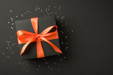 Top view photo of stylish black giftbox with orange satin ribbon bow and bright sequins on isolated black background with copyspace - obrazy, fototapety, plakaty