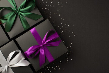 Top view photo of three stylish black gift boxes with purple green and white satin ribbon bows and sequins on isolated black background with empty space